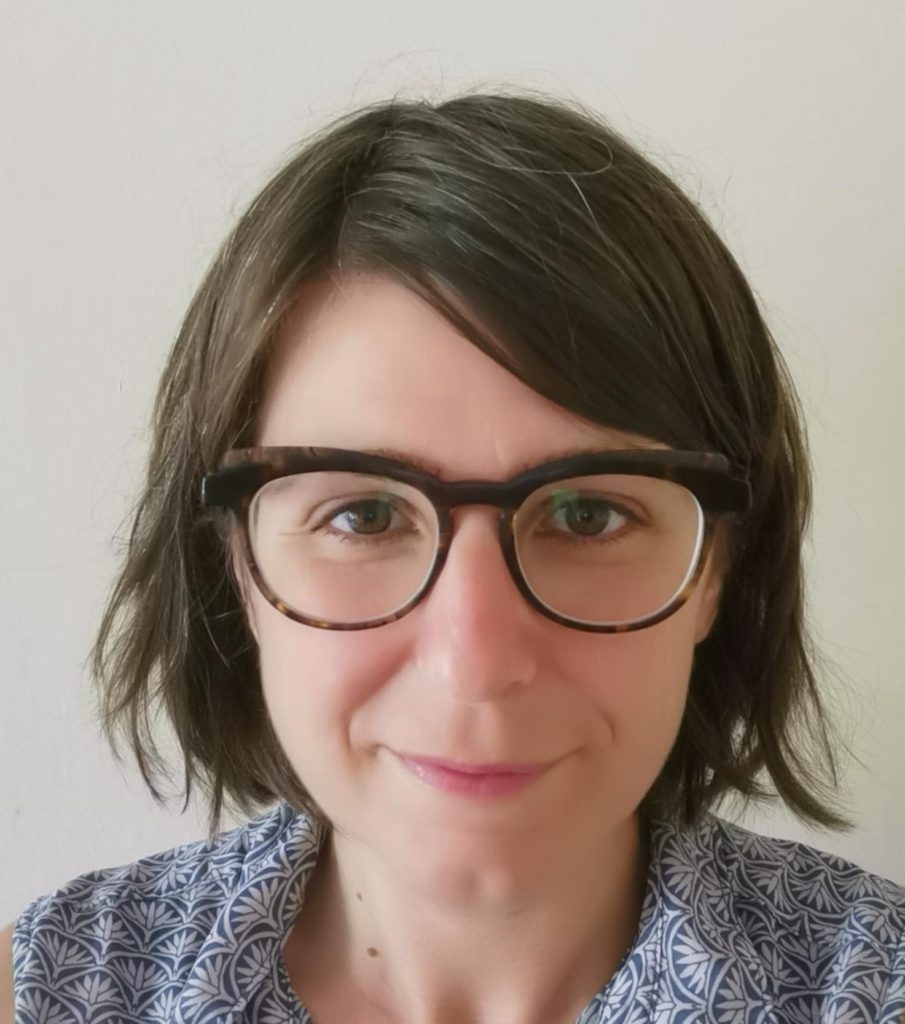 Person with short brown hair and glasses, wearing a patterned shirt, smiles at the camera against a plain background.