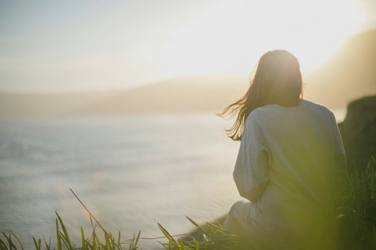 Person sitting on grass, facing a body of water, with sunlight creating a warm glow.