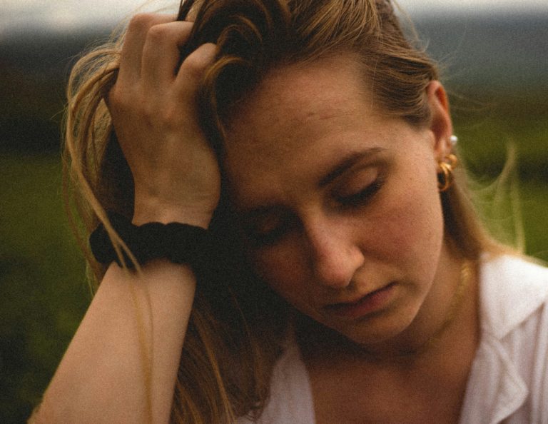 A person with long hair rests their head on their hand, standing outdoors.