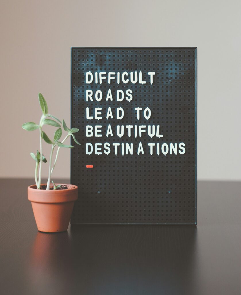 Sign with the message "DIFFICULT ROADS LEAD TO BEAUTIFUL DESTINATIONS" next to a small potted plant on a table.