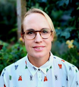 A person with short blonde hair and glasses, wearing a white shirt with colorful insect patterns, stands outdoors with leafy greenery in the background.
