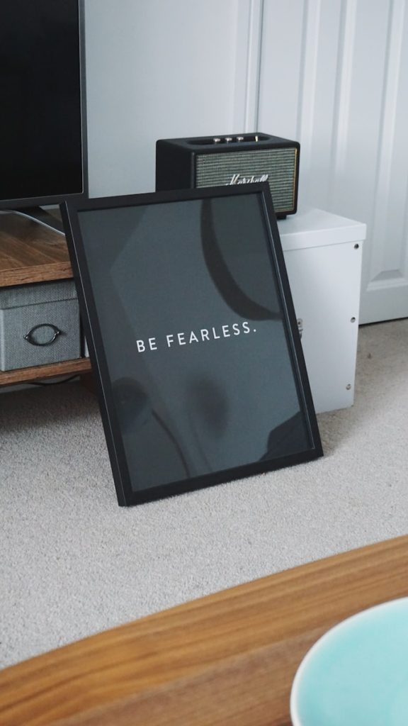 Framed sign with "BE FEARLESS." leaning against furniture, near a small speaker and a white cabinet on a carpeted floor.