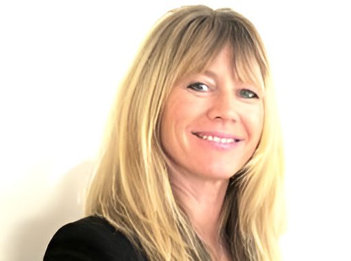 A woman with long blonde hair smiles, wearing a dark top, against a plain background.