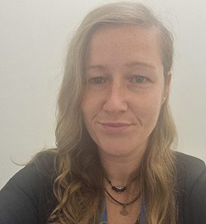 Person with long hair wearing a black top and necklaces, standing in front of a plain white background, looking at the camera.