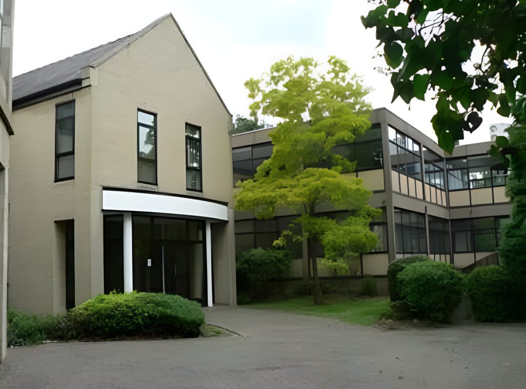 Office building with beige walls and large windows, surrounded by greenery and a prominent tree in the courtyard.