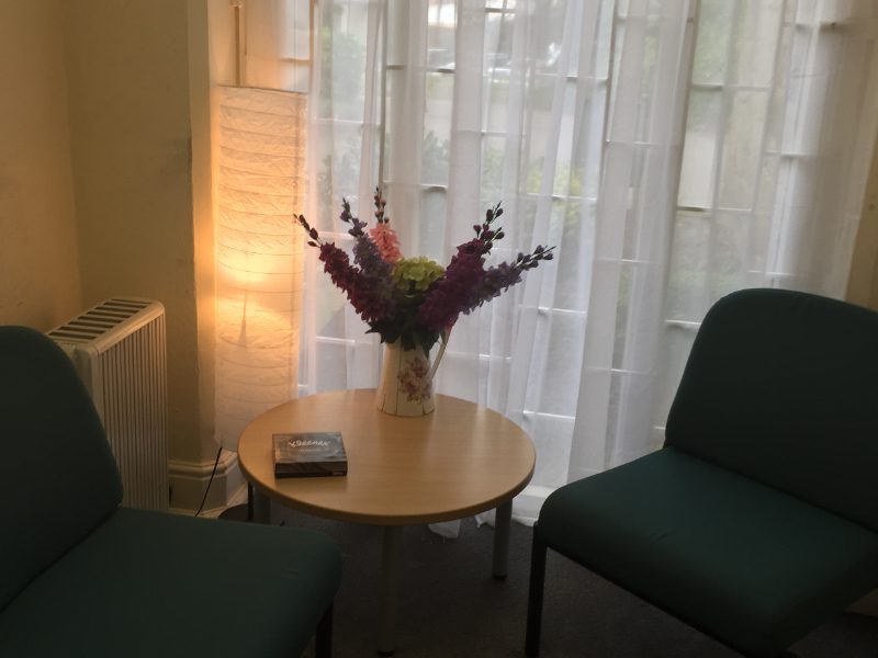 Two green chairs face a round wooden table with a floral vase and a book. A tall lamp stands beside a radiator, near sheer curtains.