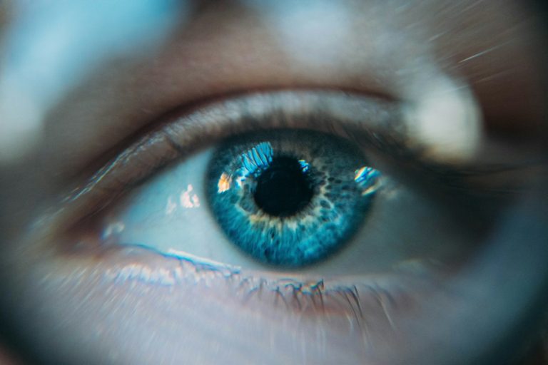 Close-up of a blue eye showing detailed iris patterns and reflections, with focus on the pupil and surrounding textures.