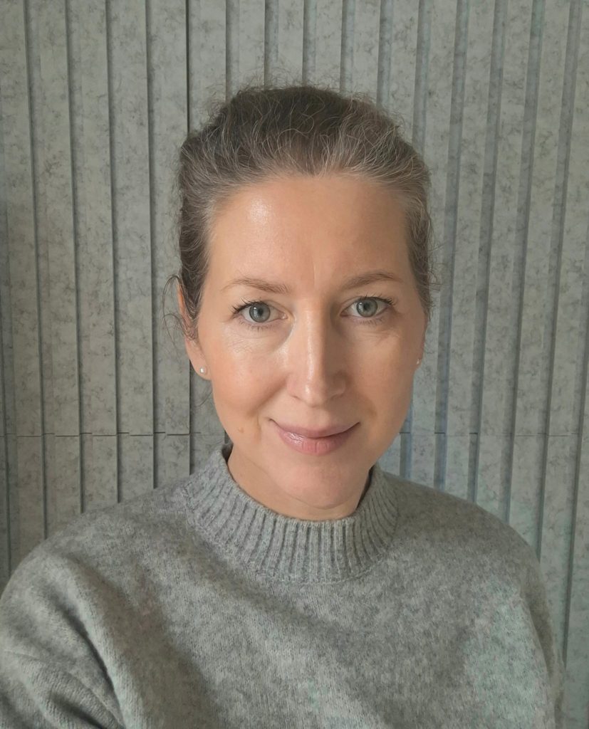 A person with tied-back hair and a grey sweater smiles slightly in front of a vertically striped background.
