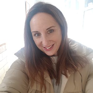 Woman with long brown hair, wearing a beige puffer jacket, in a well-lit room.