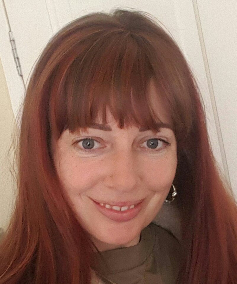 A woman with long reddish-brown hair and bangs smiles at the camera, standing indoors in front of a white door.
